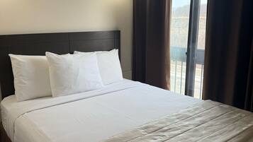 A neatly made bed with white linens and a beige patterned throw, a dark headboard, and a window with curtains.