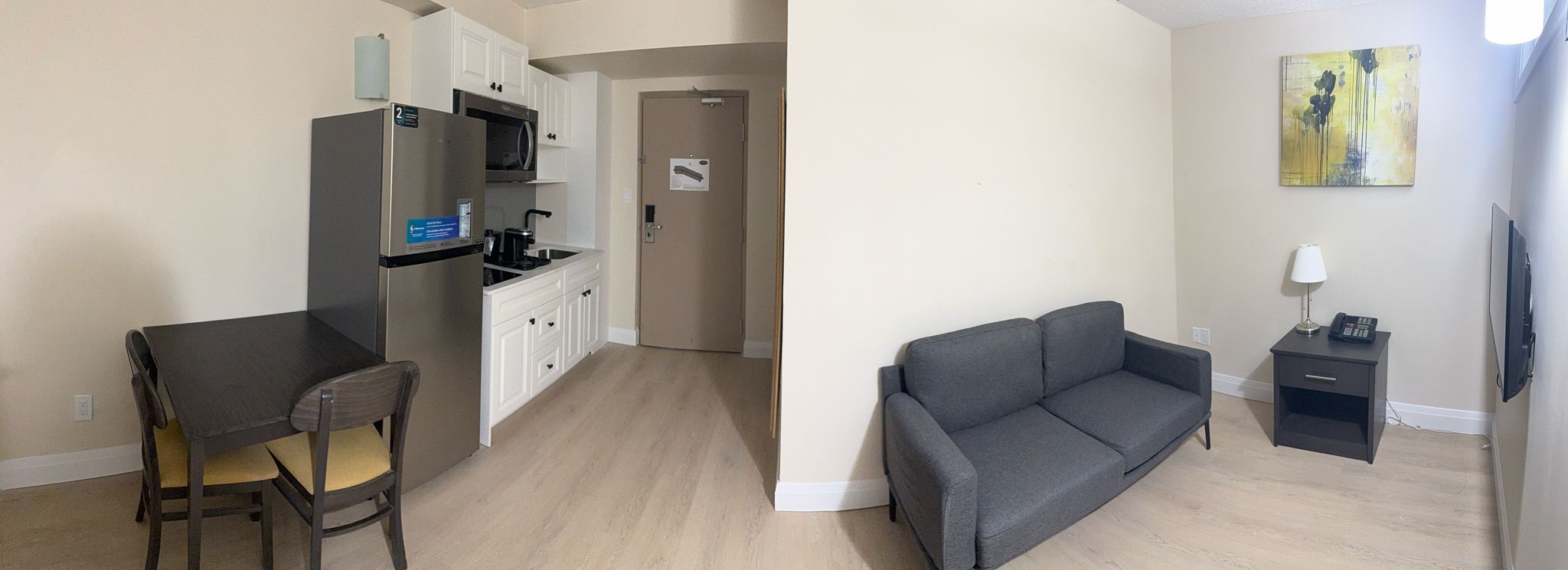 A compact living space with a kitchenette, a dining table with a chair, a gray sofa, a small side table, and a painting on the wall.