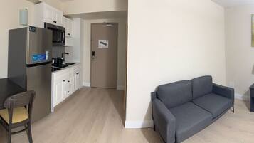 A compact living space with a kitchenette, a dining table with a chair, a gray sofa, a small side table, and a painting on the wall.