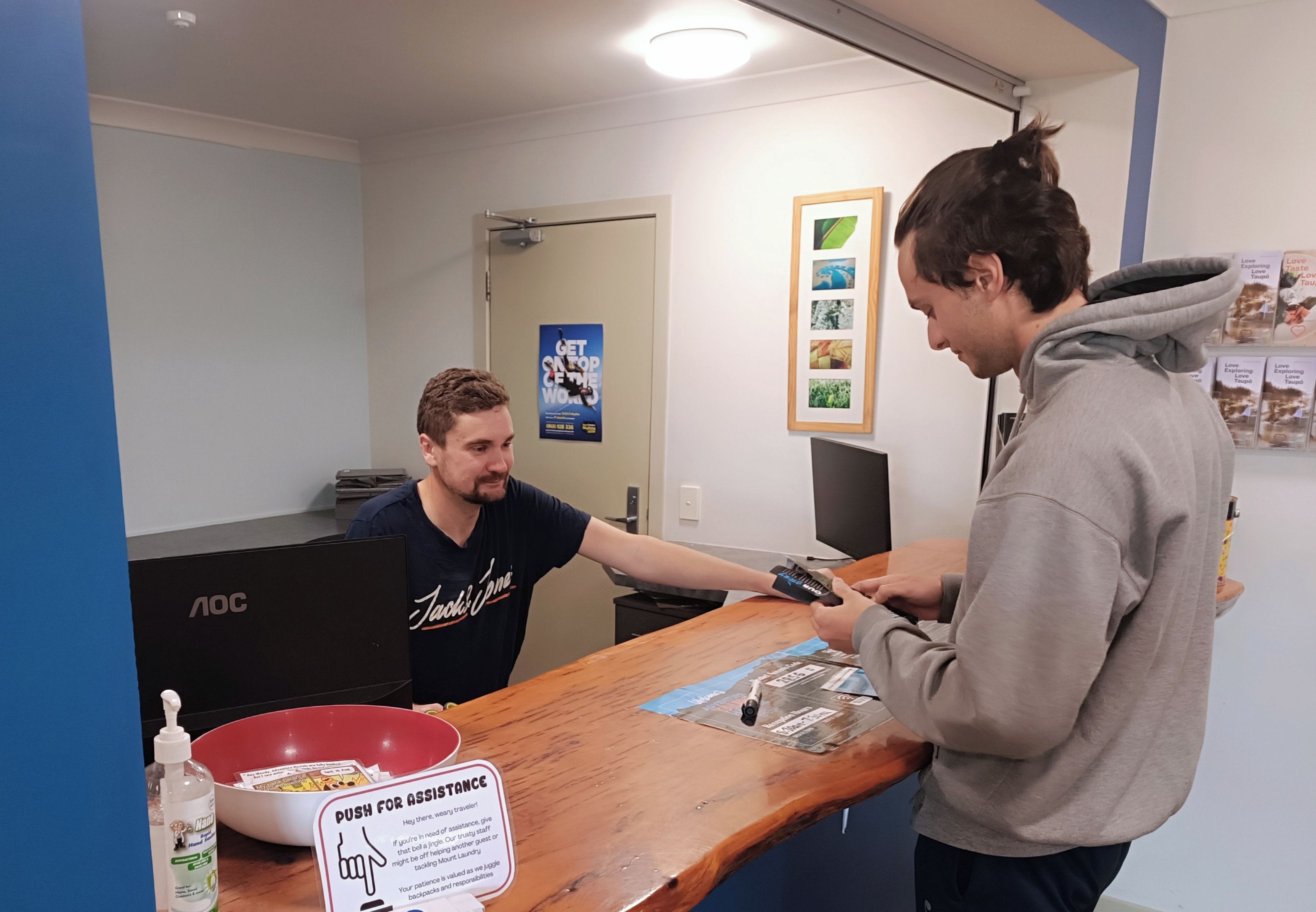 A person handing over a card to another person at a counter with a sign that reads 'PUSH FOR ASSISTANCE'.