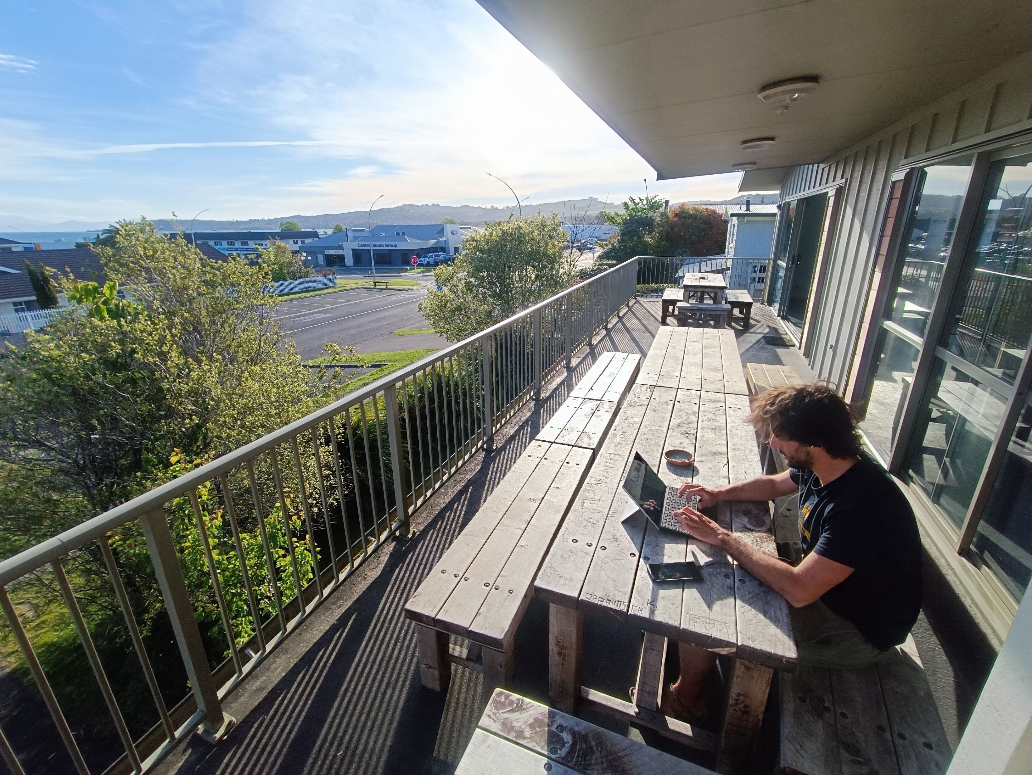 A balcony with a view of a road and buildings, a wooden table, and a person using a laptop.