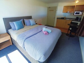 A hotel room with a bed, a bedside table, a small desk, a microwave, and a kitchenette.