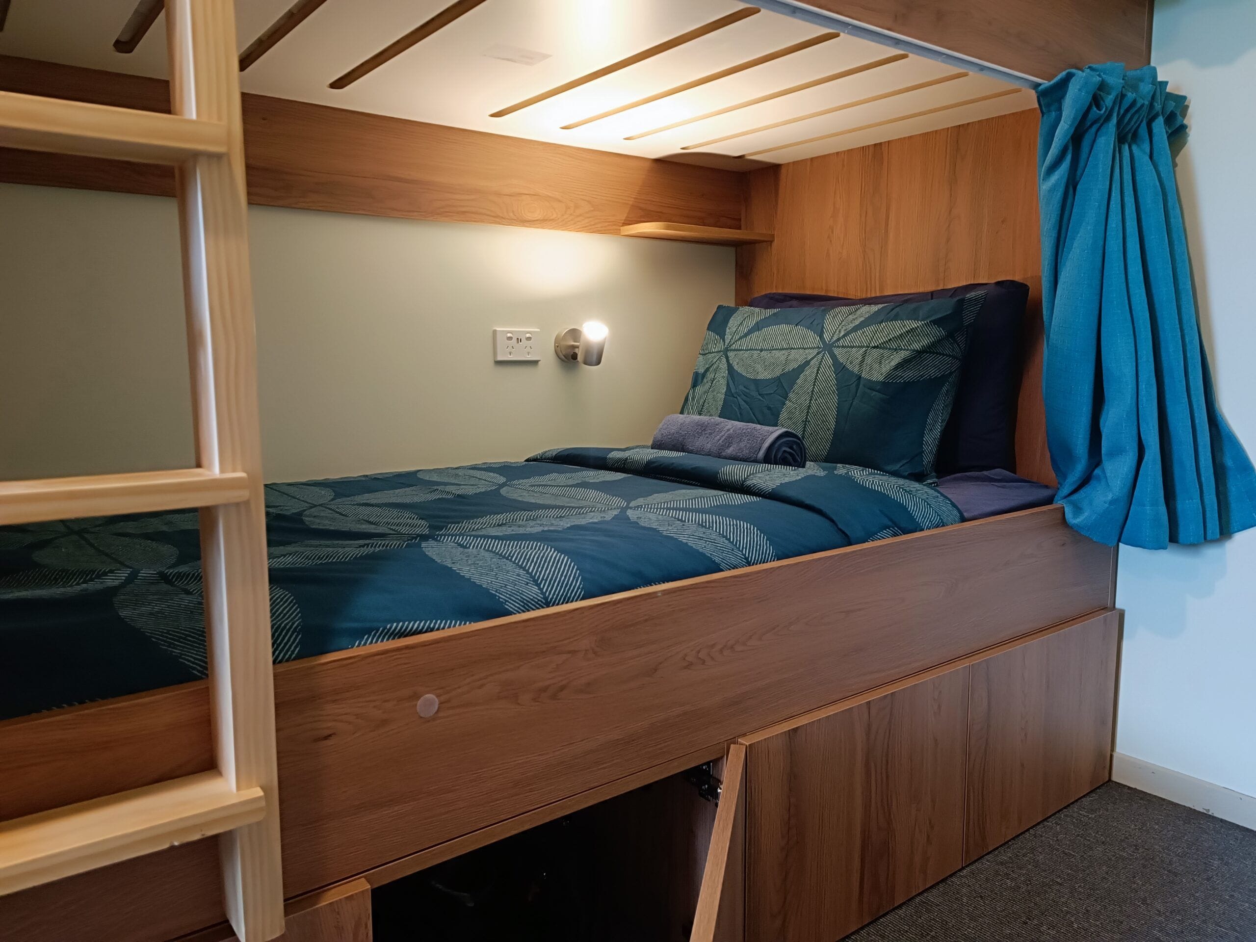 Quarto compacto com uma cama beliche, uma mesinha de cabeceira, uma luminária na parede e uma cortina.