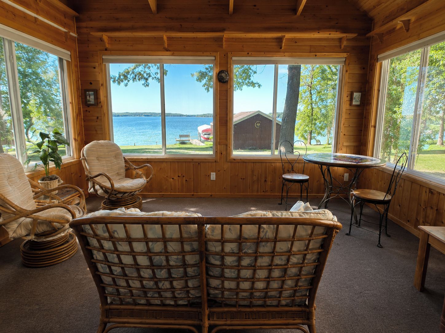 A cozy cabin interior with wicker furniture, large windows offering a view of a lake and dock, and a wooden ceiling.