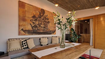 Een moderne woonkamer met een houten eettafel, een grijze bank met decoratieve kussens, een groot abstract schilderij en een vaas met bloemen.