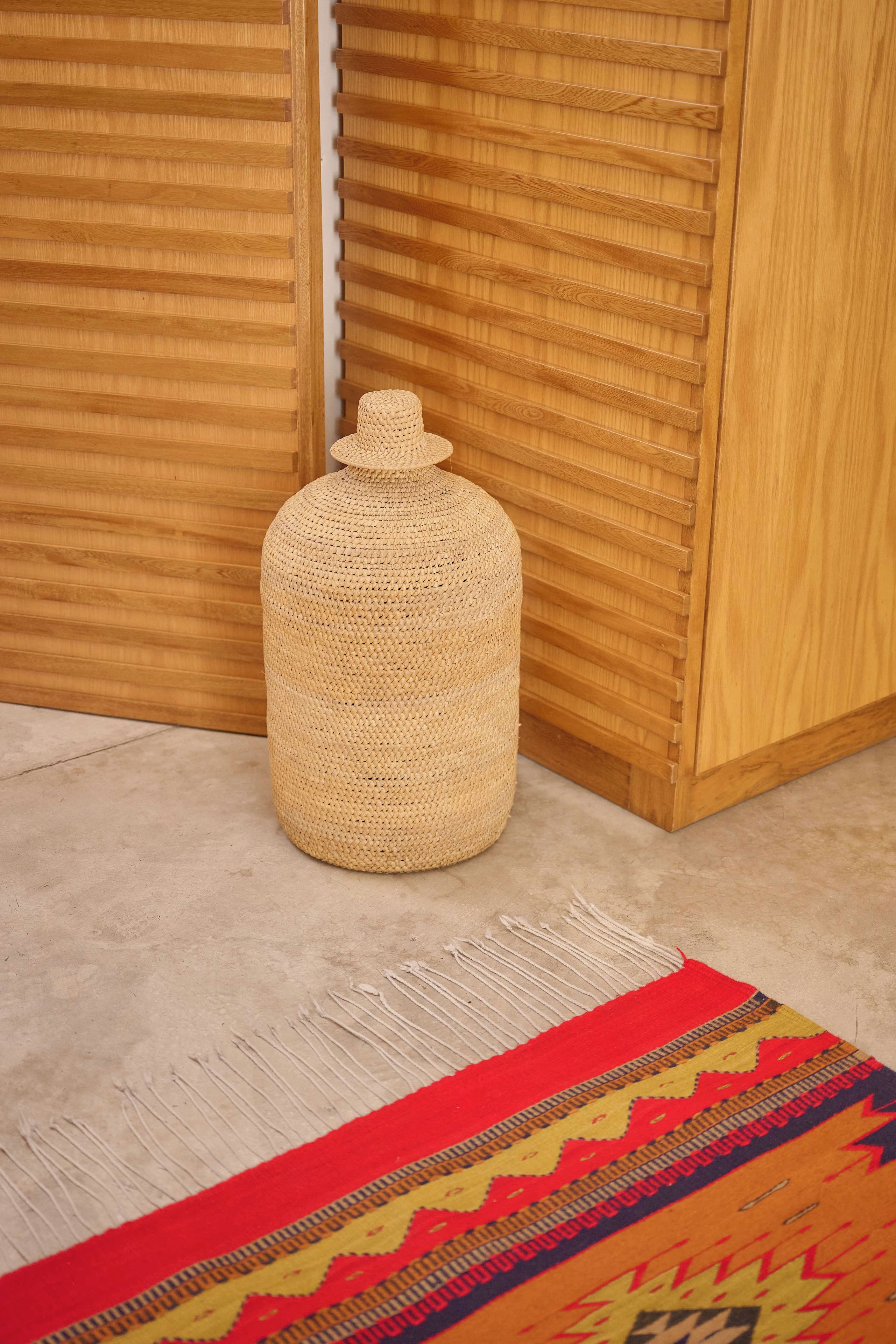 Un panier tressé, placé dans un coin d’une pièce aux murs lambrissés et ornée d’un tapis à motifs.