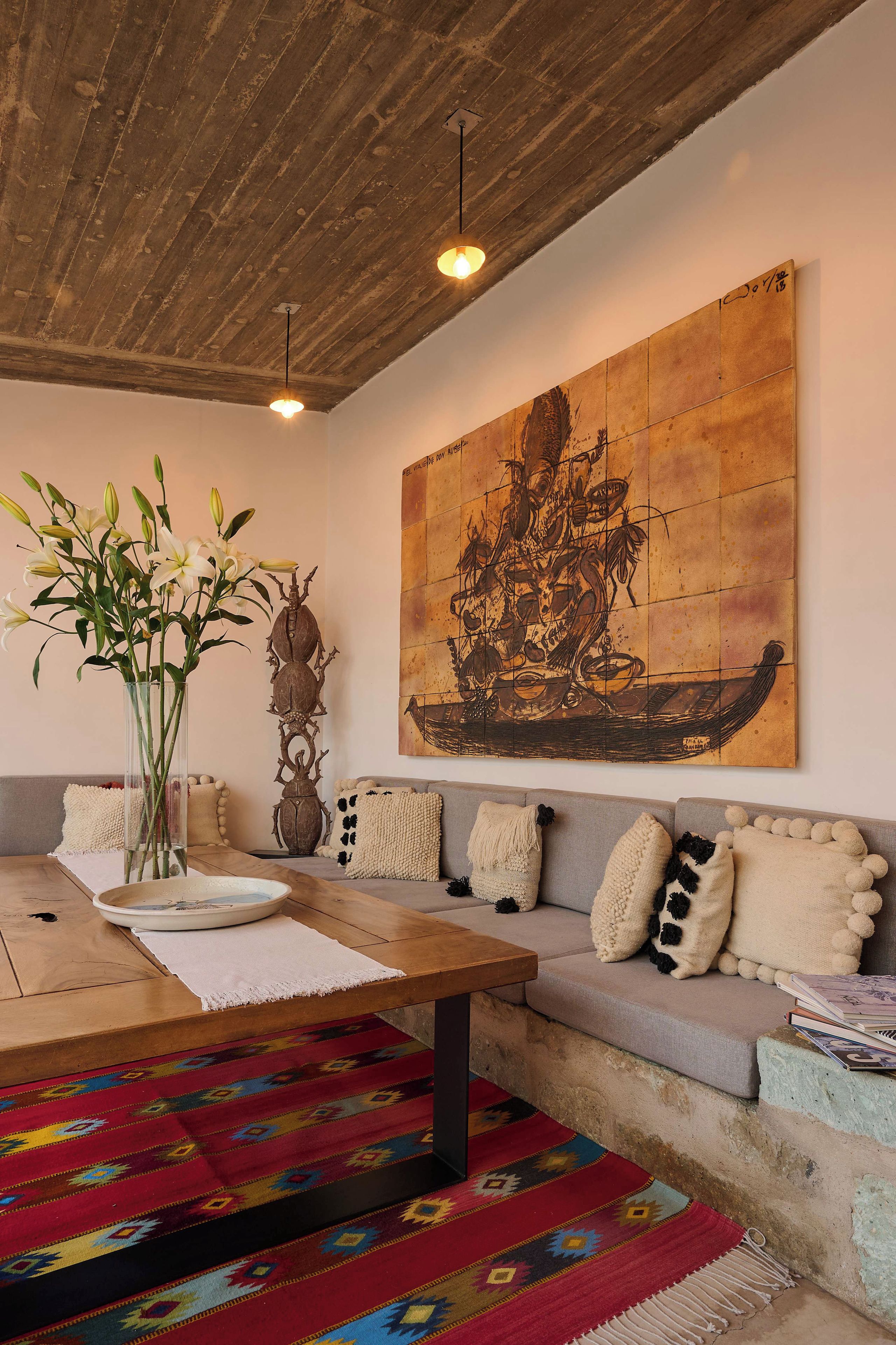 A living room with a wooden ceiling, a large abstract painting, a sofa with decorative pillows, a coffee table with a vase of flowers, and a colorful rug.