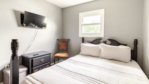 Un dormitorio con cama, cómoda, silla, televisor montado en la pared y ventana.