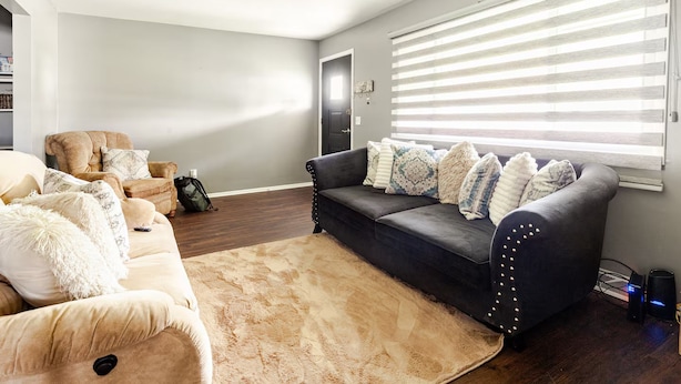 Una sala de estar con un sofá color beige, un sofá gris oscuro con cojines estampados, una mesa de centro de madera y un ventanal con persianas.