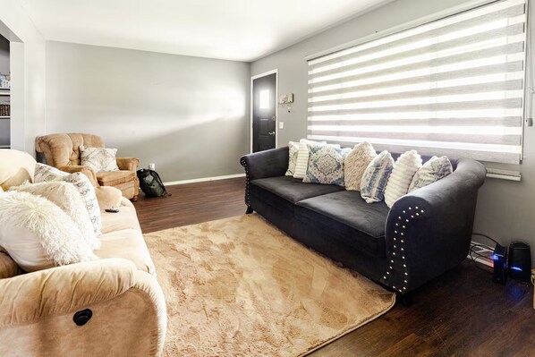 A living room with a beige sofa, a dark grey sofa with patterned cushions, a wooden coffee table, and a large window with blinds.