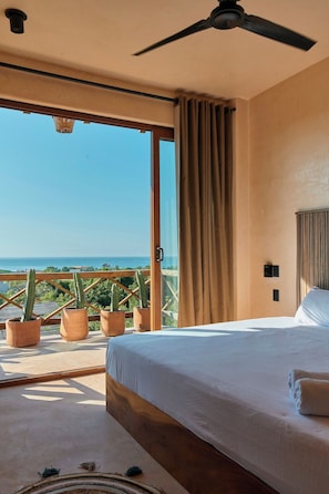 Een slaapkamer met een bed, een plafondventilator, uitzicht op de oceaan en potplanten met cactussen.