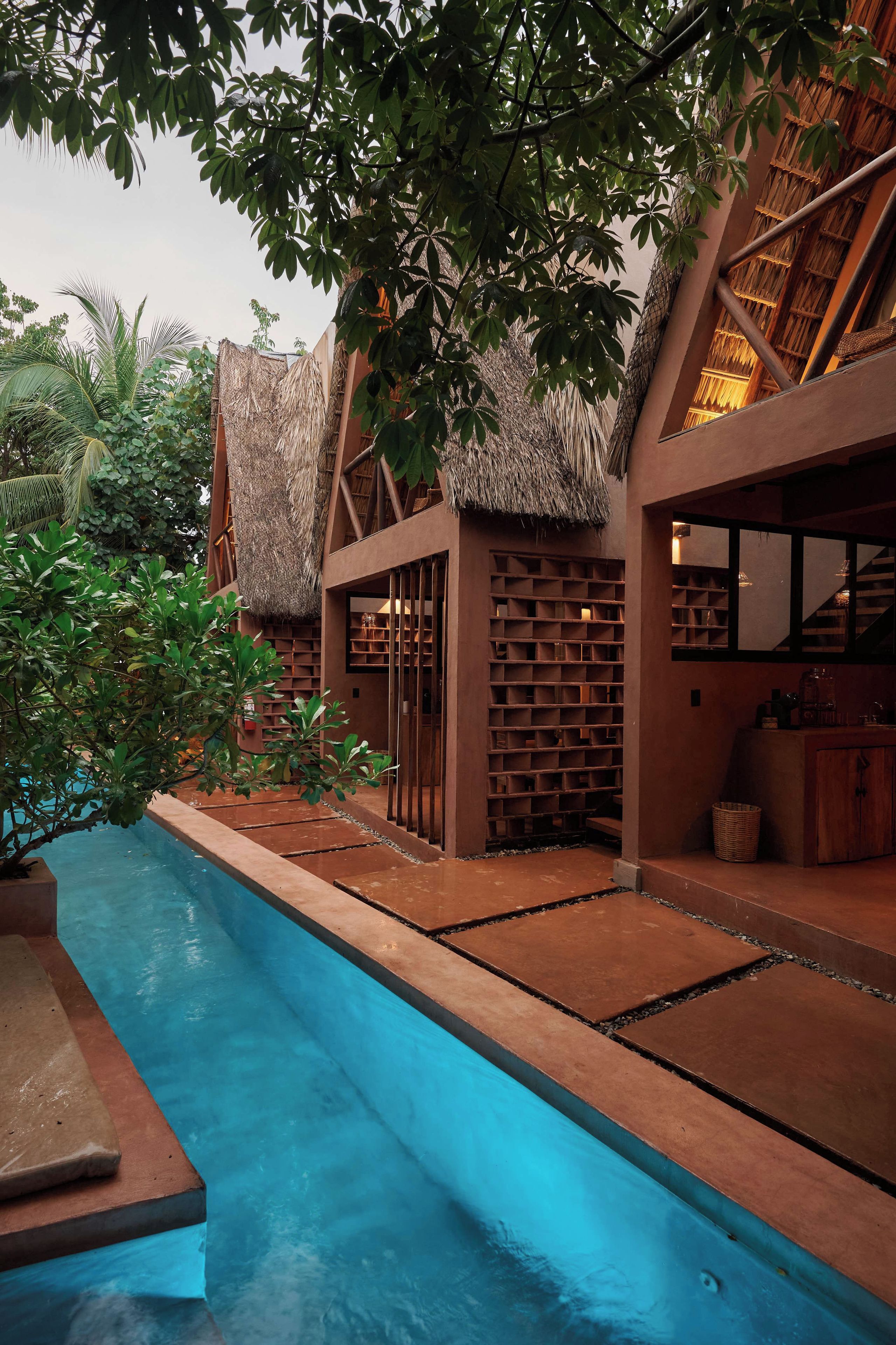 A pool area with a thatched roof structure, surrounded by greenery and a modern building with a brick facade.