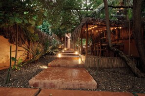 A pathway leading to a building with a thatched roof and string lights, surrounded by greenery.