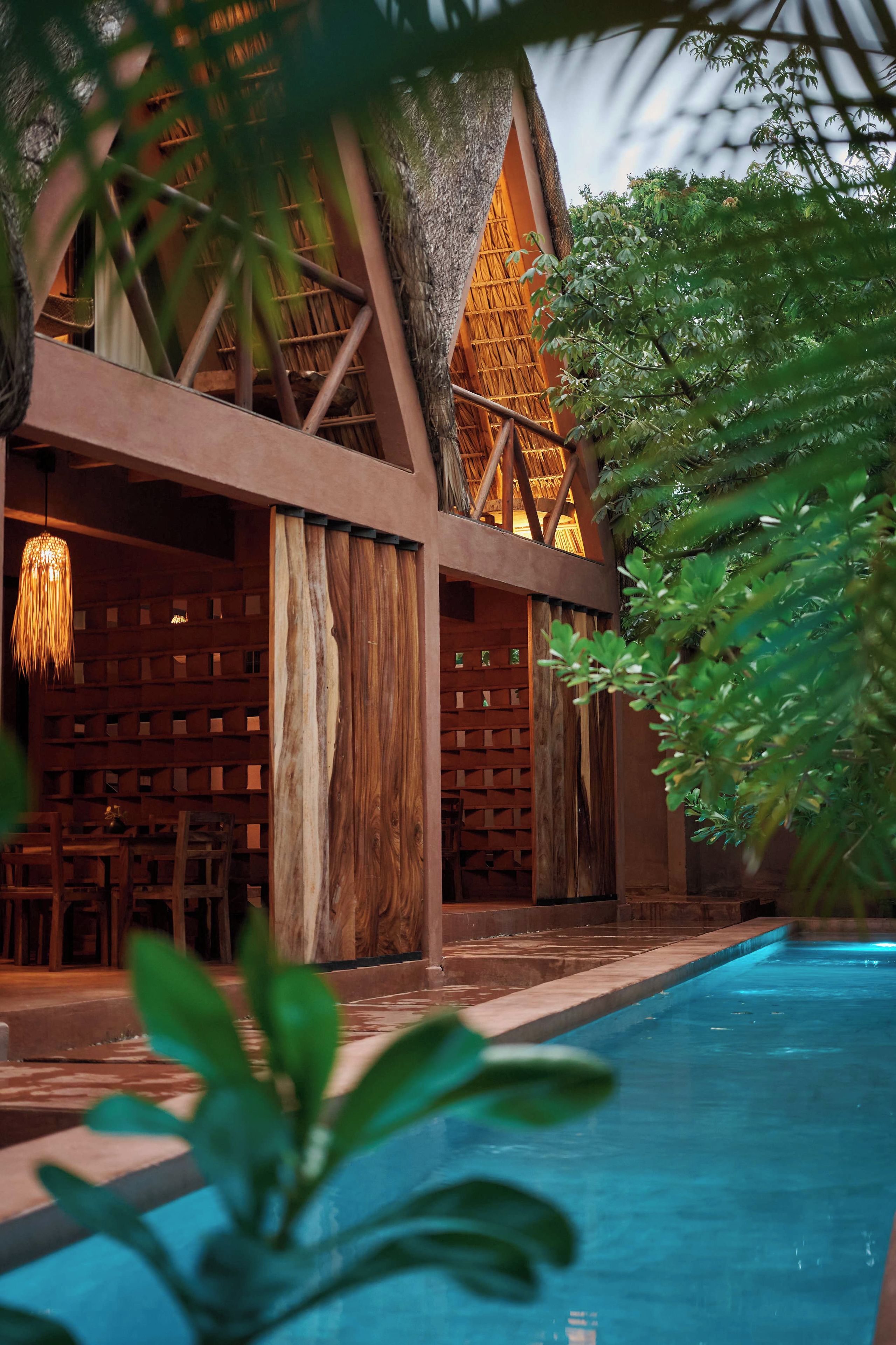 Área da piscina com estrutura de madeira, luminária pendente e piscina azul cristalina.