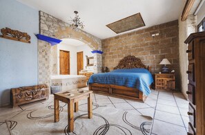 Un dormitorio con pared de ladrillo, una cama con cubrecama azul, un banco de madera, una mesita y un espejo en la pared.