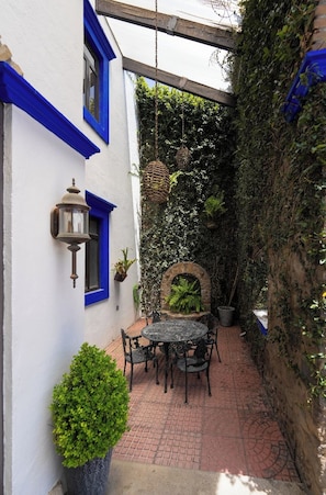 Une terrasse avec une table et des chaises, entourée d’un mur blanc orné de bordures bleues, d’une jardinière suspendue et de plantes en pot.