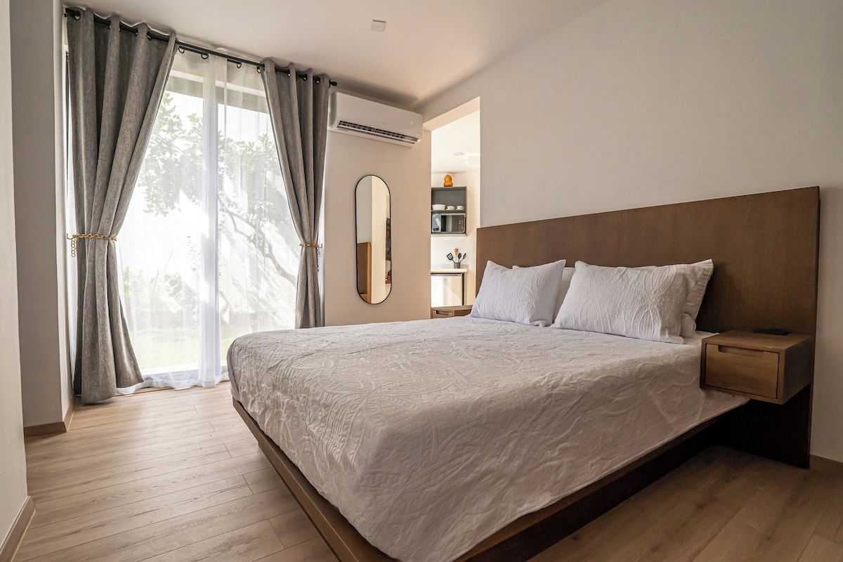 Ein Schlafzimmer mit einem großen Bett, einem Holzkopfende, einem Nachttisch, einem Spiegel und Blick ins Freie durch transparente Vorhänge.