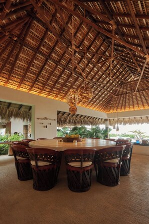 A spacious dining area with a thatched roof and a large round table surrounded by chairs. - Private beachfront estate in the Oaxacan jungle (Puerto Escondido)