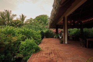 A brick pathway leading to a covered outdoor area with a thatched roof, surrounded by lush greenery and palm trees.
