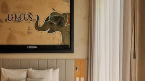 Una camera da letto con un'ampia opera d'arte incorniciata raffigurante un elefante, un letto con numerosi cuscini, un comodino e una finestra con tende.