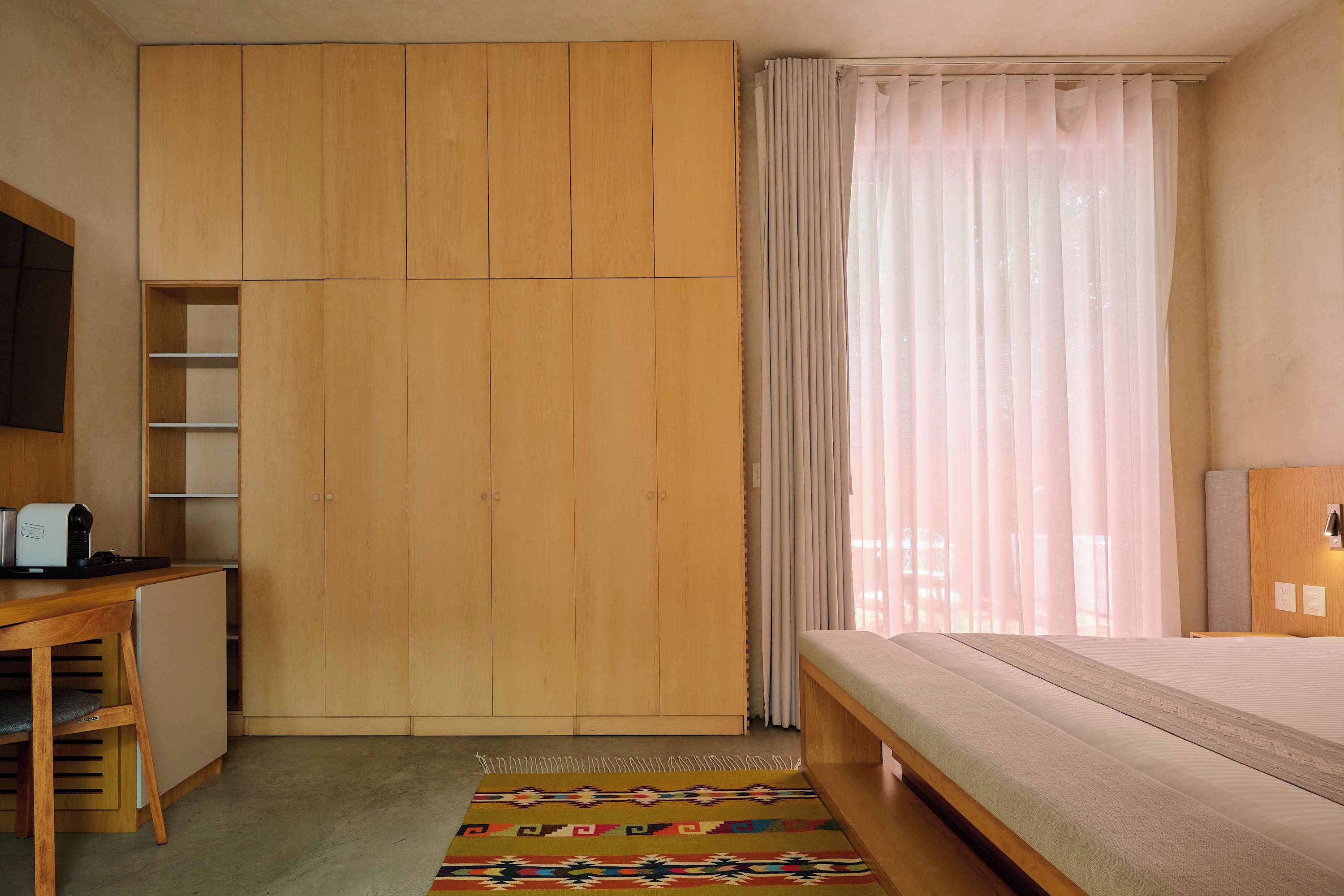 A bedroom with a bed, a wooden wardrobe, a desk with a chair, and a window with curtains.