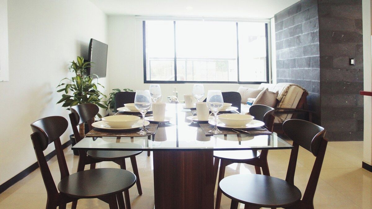 A dining area with a large table set for six, surrounded by chairs, with a plant and a TV on the wall.
