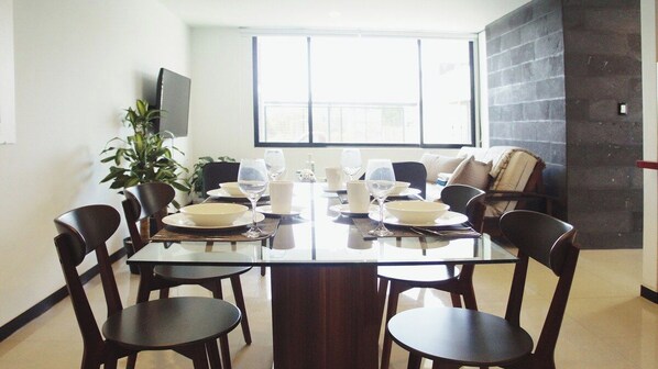 A dining area with a large table set for six, surrounded by chairs, with a plant and a TV on the wall.