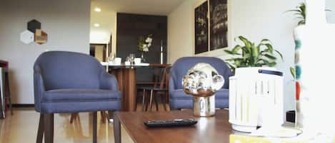 Ein Wohnzimmer mit zwei blauen Sesseln, einem Holztisch, einer Fernbedienung, einer dekorativen Metallskulptur und einem weißen Gerät rechts.