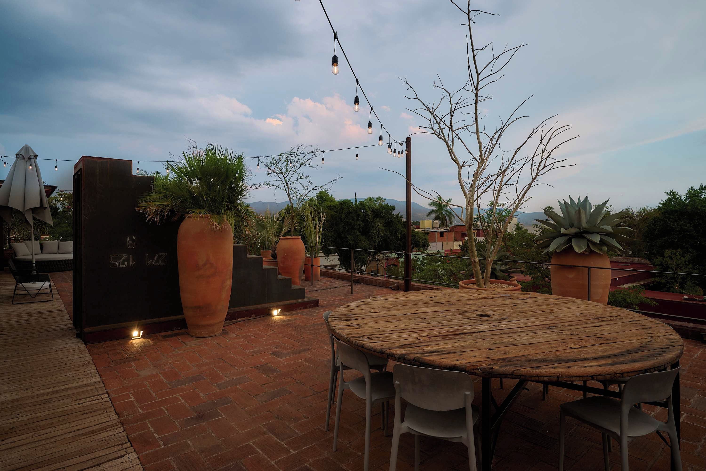 Un patio al aire libre con una mesa y sillas de madera redonda, macetas de terracota y luces colgantes.