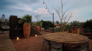 Een terras buiten met een ronde houten tafel en stoelen, terracotta plantenbakken en lichtslingers.