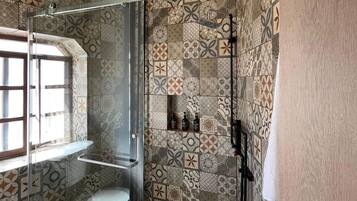 A bathroom with a glass-enclosed shower, patterned tiles, a toilet, and a window.
