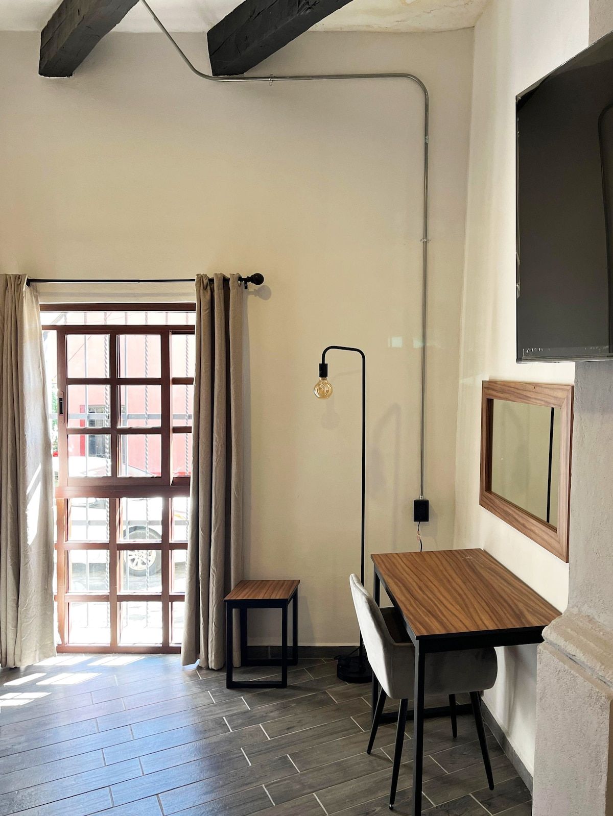 A room with a wooden desk, a chair, a small table, a floor lamp, and a large window with curtains.
