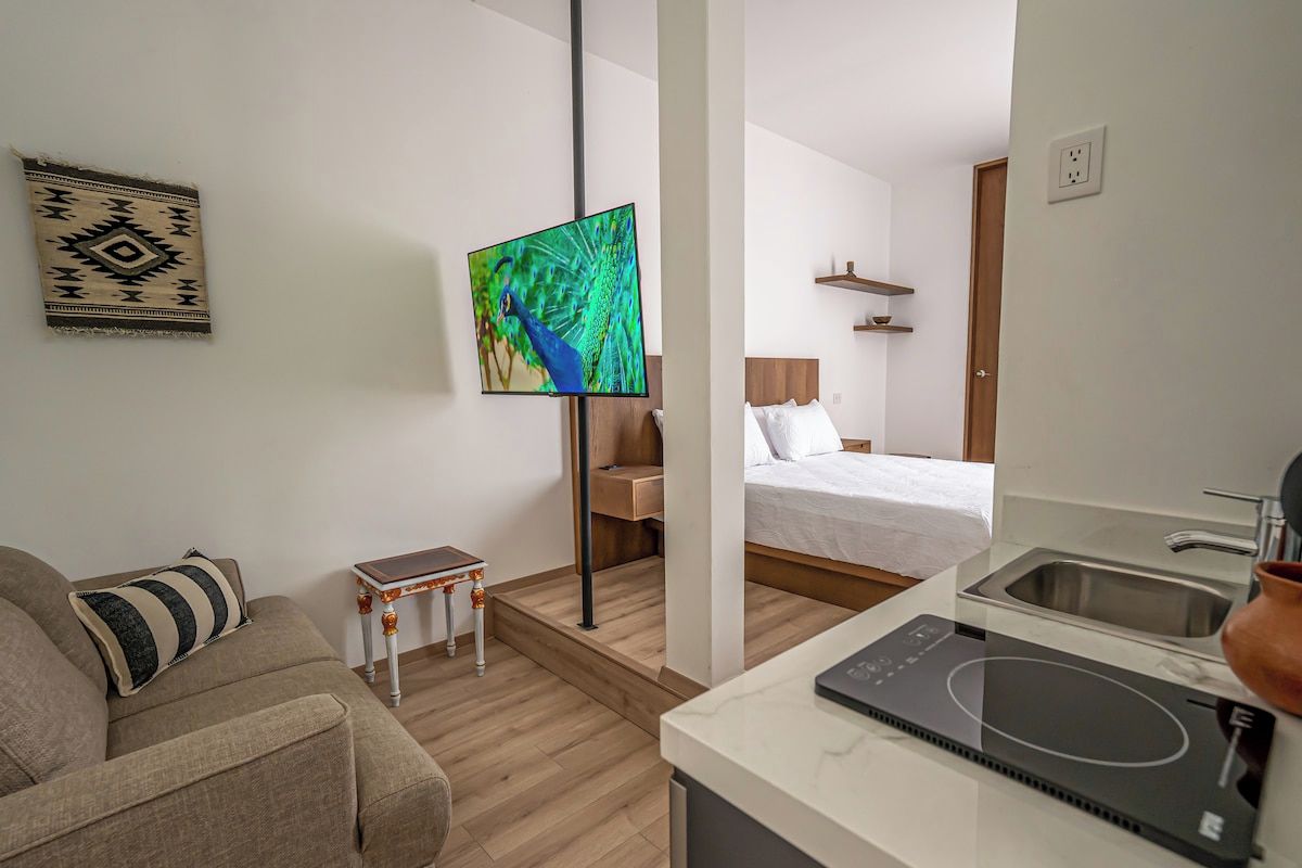 Een moderne hotelkamer met een bed, een televisie, een tafeltje en een kitchenette met een spoelbak en fornuis.