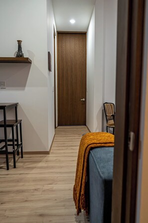 Un couloir étroit, avec une porte en bois au fond, un bureau avec une chaise et un lit recouvert d’une couverture.
