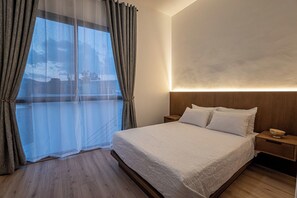 A bedroom with a large bed, a nightstand, a bedside table, and a view through a window with curtains. - Laurelian 8 · Eles · 2 BR Apt near downtown (Oaxaca de Juárez)