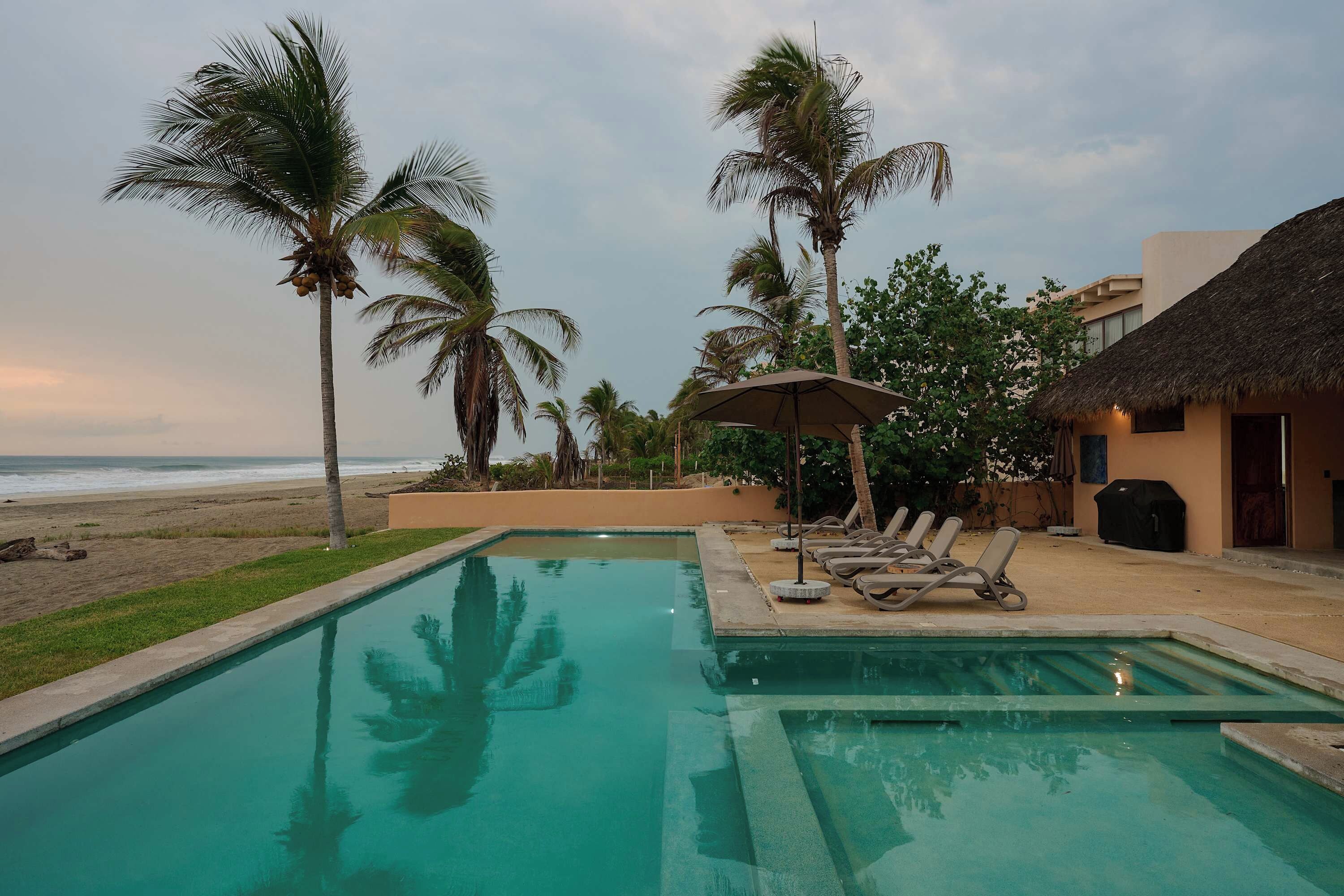 A pool area with lounge chairs and umbrellas, palm trees, and a beach view.