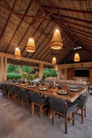 A spacious dining area with a high thatched ceiling, multiple hanging light fixtures, and a long wooden table set for a meal. - Casa Juliana · Private suite · Beachfront property (Puerto Escondido)