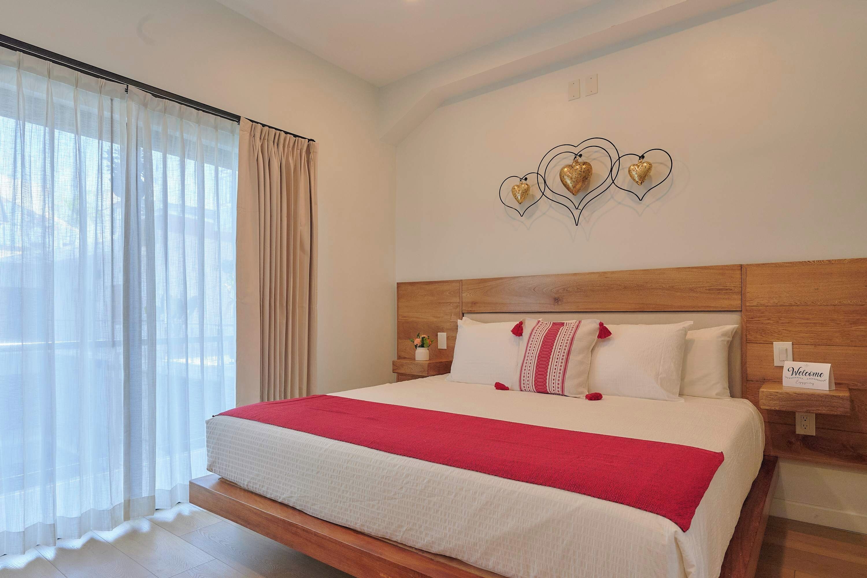 A hotel room with a large bed, a wooden headboard, a bedside table with a lamp, and a wall-mounted heart decoration.