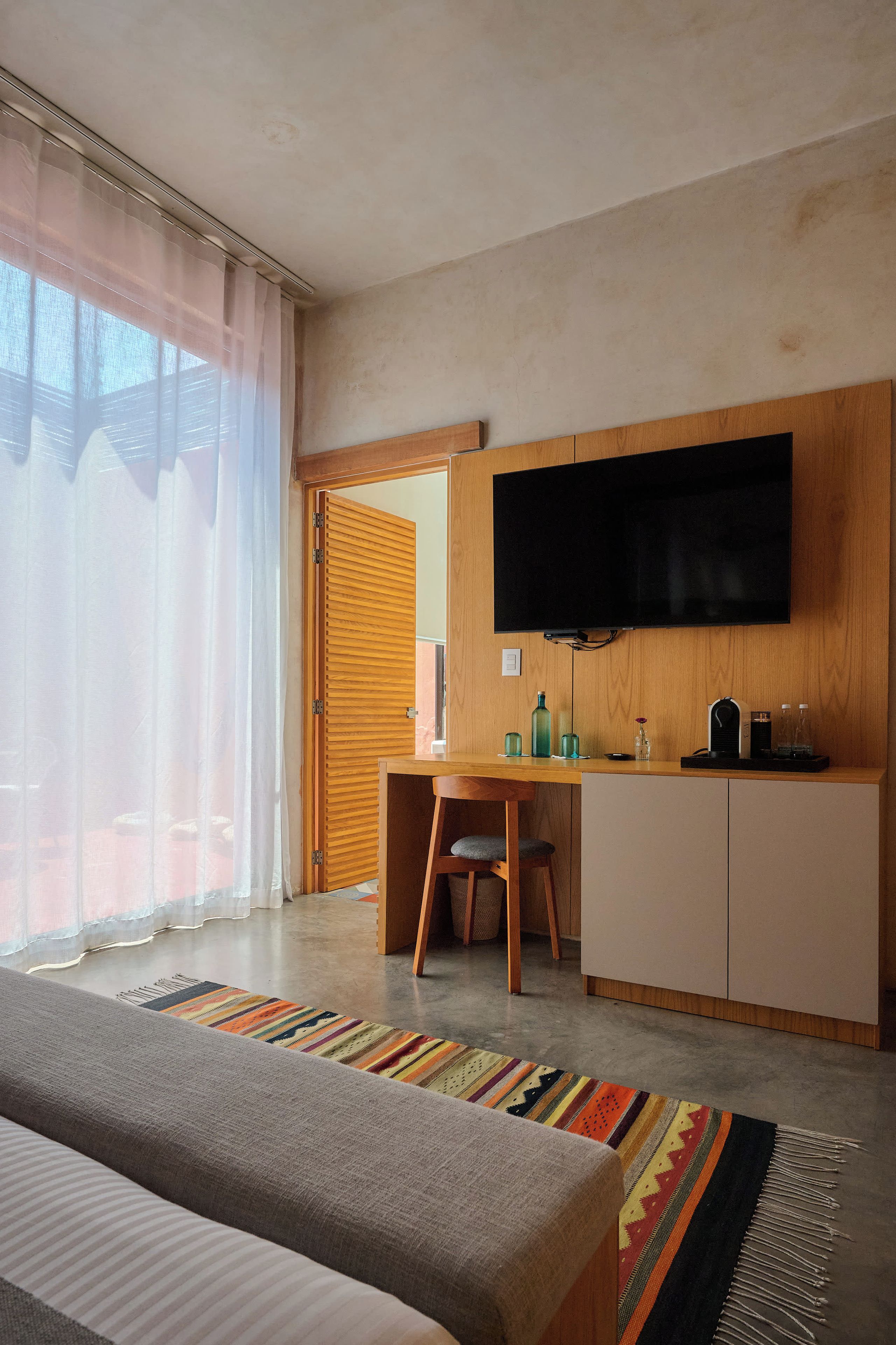 Una habitación de hotel moderna con una cama, un escritorio, un televisor y vistas al exterior a través de una ventana con cortinas translúcidas.