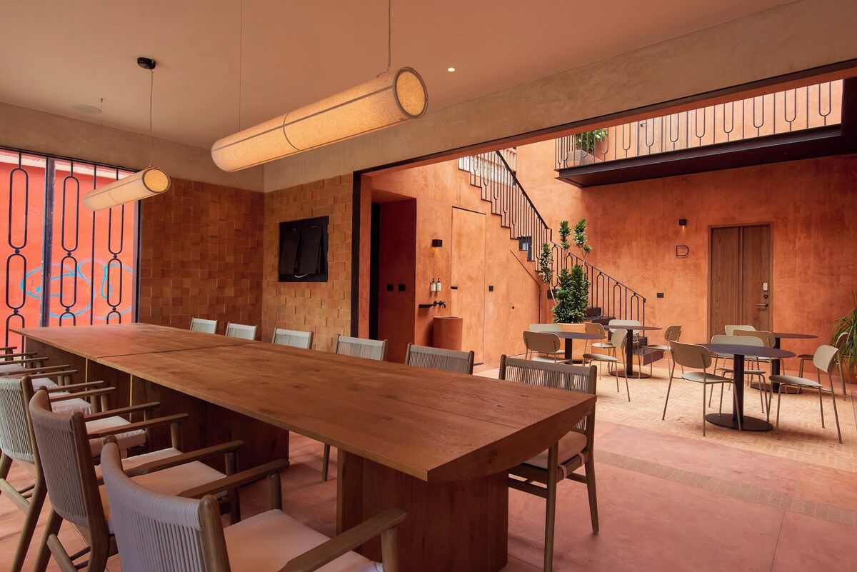 A spacious indoor area with a long wooden table and chairs, a staircase, and a balcony with plants.