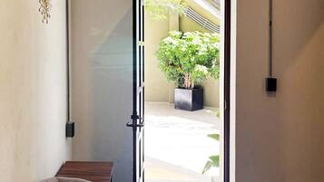 Um espaço interno com sofá, porta que dá para área externa com planta em vaso e luminária fixada na parede.
