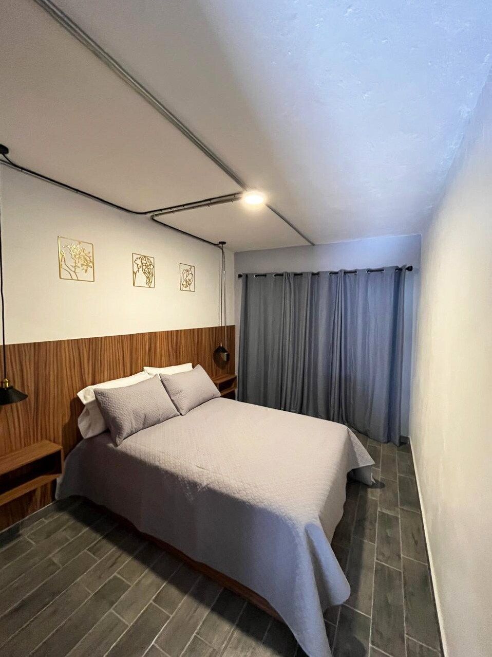 A hotel room with a bed, a wooden headboard, a nightstand, a wall-mounted lamp, and a window with curtains.