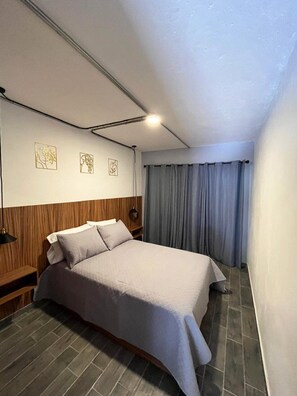 Habitación de hotel con cama, cabecero de madera, mesita de noche, lámpara de pared y ventana con cortinas.