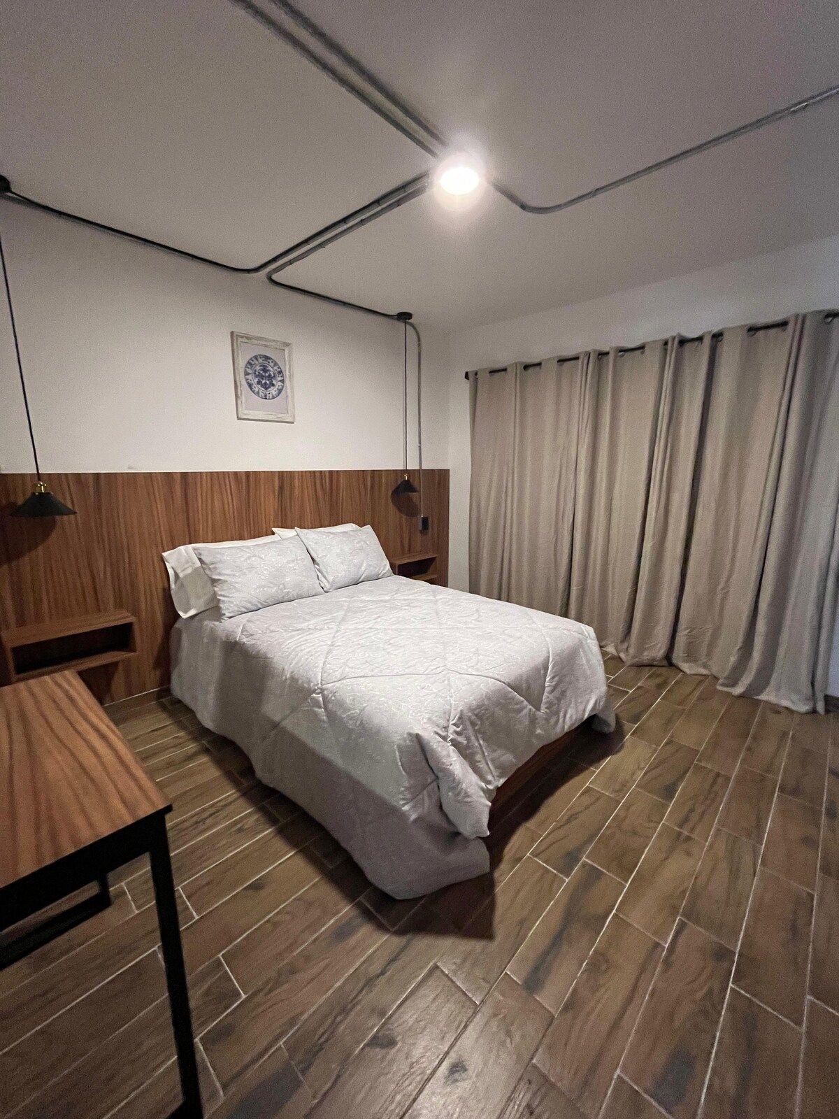 Habitación de hotel con cama, cabecera de madera, mesita de noche, reloj en la pared y ventana con cortinas.