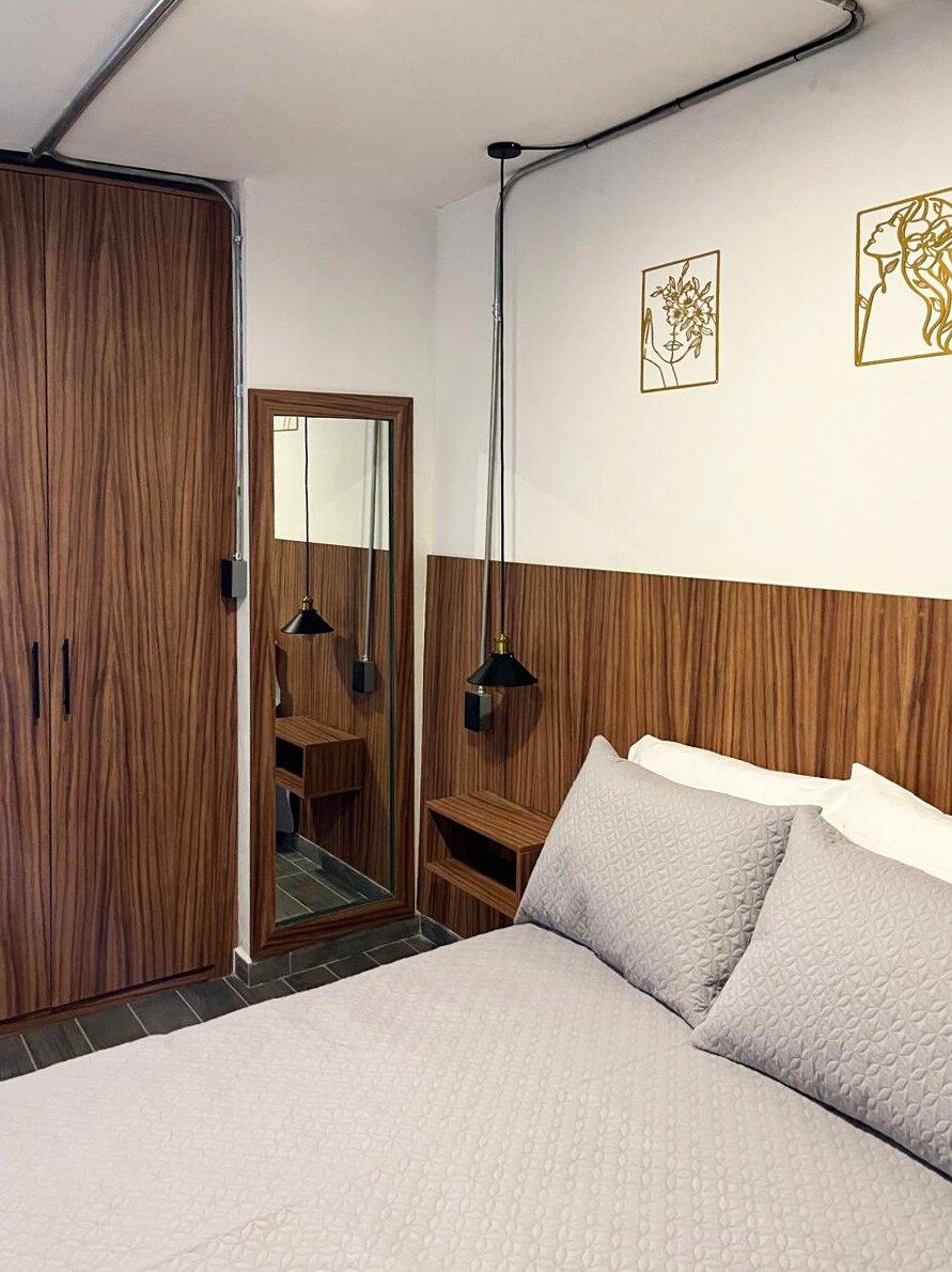 Une chambre à coucher comprenant un lit, une armoire en bois, un miroir et deux lampes fixées au mur.