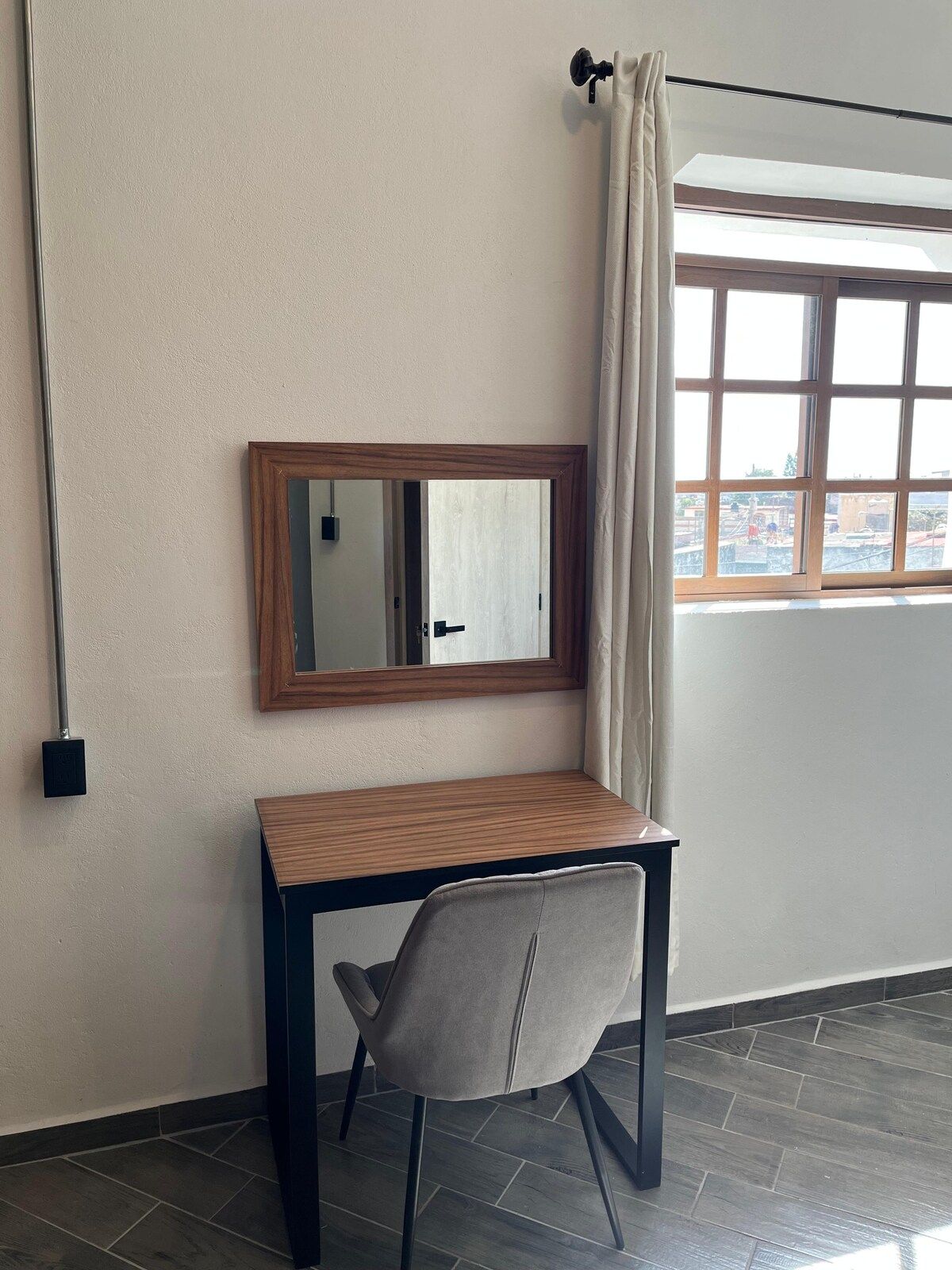 A room with a wooden desk, a chair, a mirror, a window with curtains, and a view of buildings outside.