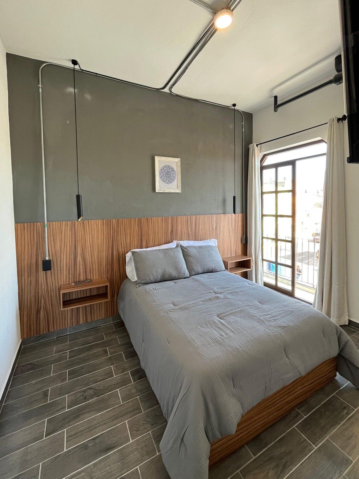Ein modernes Schlafzimmer mit einem Bett, einem Holz-Kopfteil, einer Wandleuchte und einem Fenster mit Vorhängen.