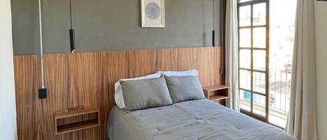 A modern bedroom with a bed, a wooden headboard, a wall-mounted light fixture, and a window with curtains.