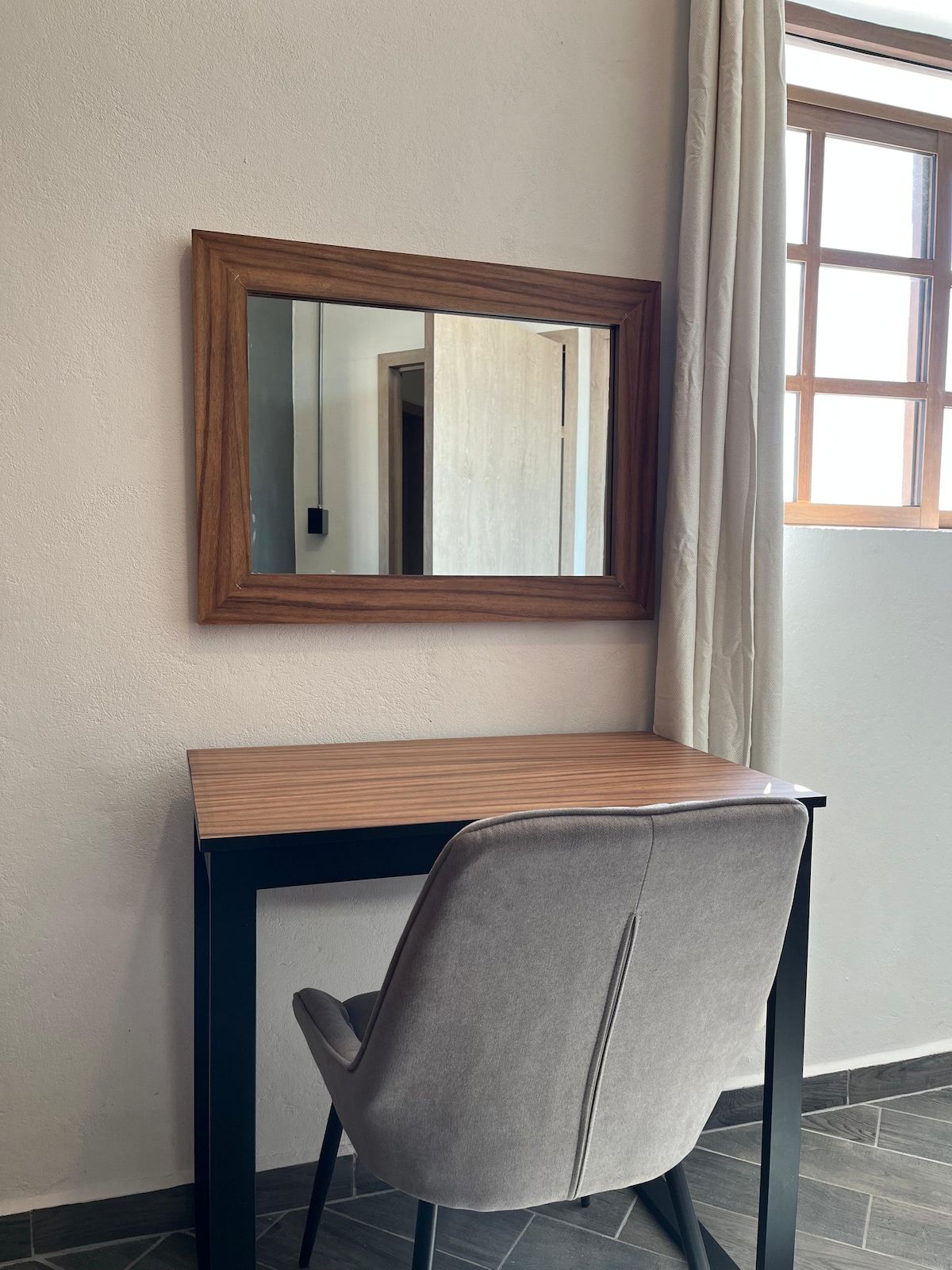A room with a wooden desk, a grey chair, a mirror, and a window with curtains.