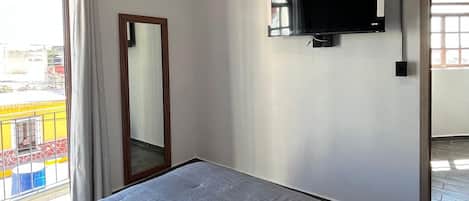 A bedroom with a bed, a flat-screen TV mounted on the wall, a window with curtains, and a view of the outside.
